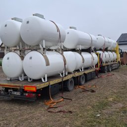 MP WORLD SPÓŁKA Z OGRANICZONĄ ODPOWIEDZIALNOŚCIĄ - Transport białych zbiorników gazowych na naczepie ciężarówki, zabezpieczonych pasami. Widok z tyłu pojazdu na tle zabudowań.