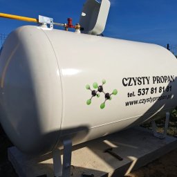 MP WORLD SPÓŁKA Z OGRANICZONĄ ODPOWIEDZIALNOŚCIĄ - Biały zbiornik na propan z żółtym przyłączem, umieszczony na betonowej podstawie. Widoczne narzędzia i logo 'Czysty Propan' z numerem telefonu i adresem strony.