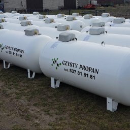 MP WORLD SPÓŁKA Z OGRANICZONĄ ODPOWIEDZIALNOŚCIĄ - Białe zbiorniki na propan z napisem 'Czysty Propan' i numerem telefonu, ustawione rzędami na zewnątrz, z budynkiem i drzewami w tle. Widok z perspektywy.