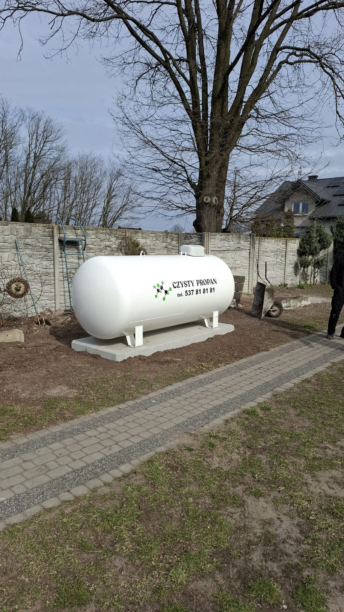 Biały zbiornik na gaz propan z napisem 'Czysty Propan' na betonowej podstawie, na tle trawnika, ogrodzenia i domu z panelami słonecznymi.