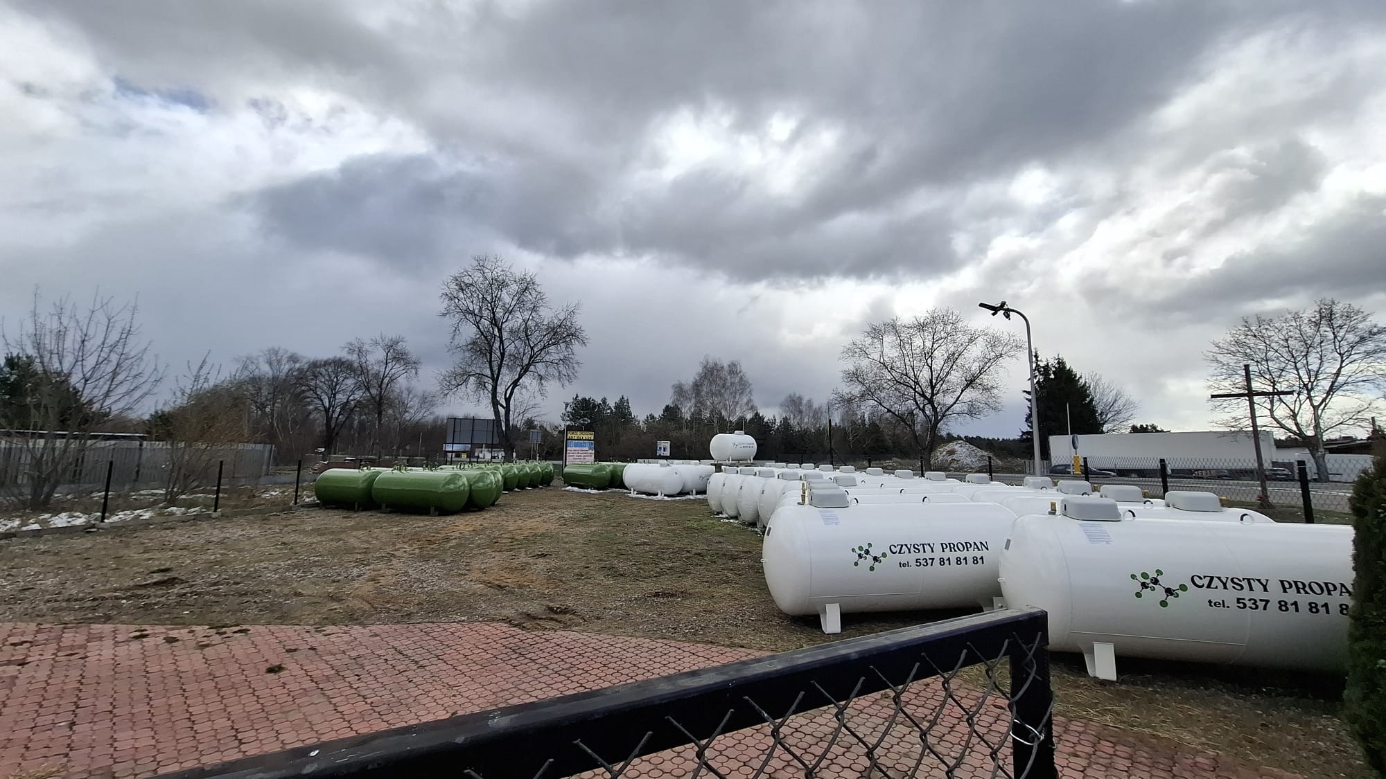 Magazyn zbiorników na gaz propan z logo firmy 'Czysty Propan' na tle pochmurnego nieba. Część zbiorników jest koloru zielonego, a część białego.