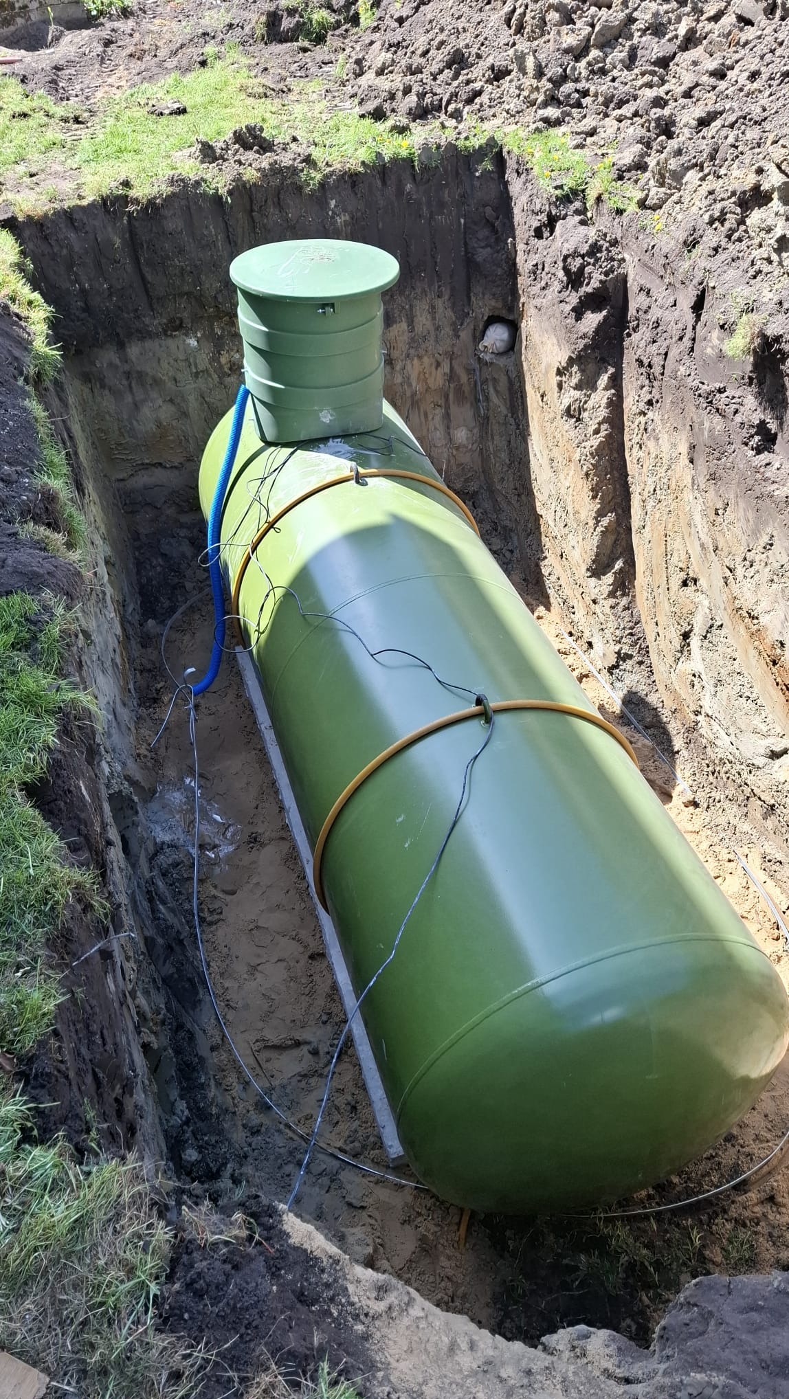 Zielony zbiornik na gaz LPG umieszczony w wykopie, zabezpieczony pasami i podłączony niebieskim przewodem. Widoczne elementy instalacji i ziemny wykop.