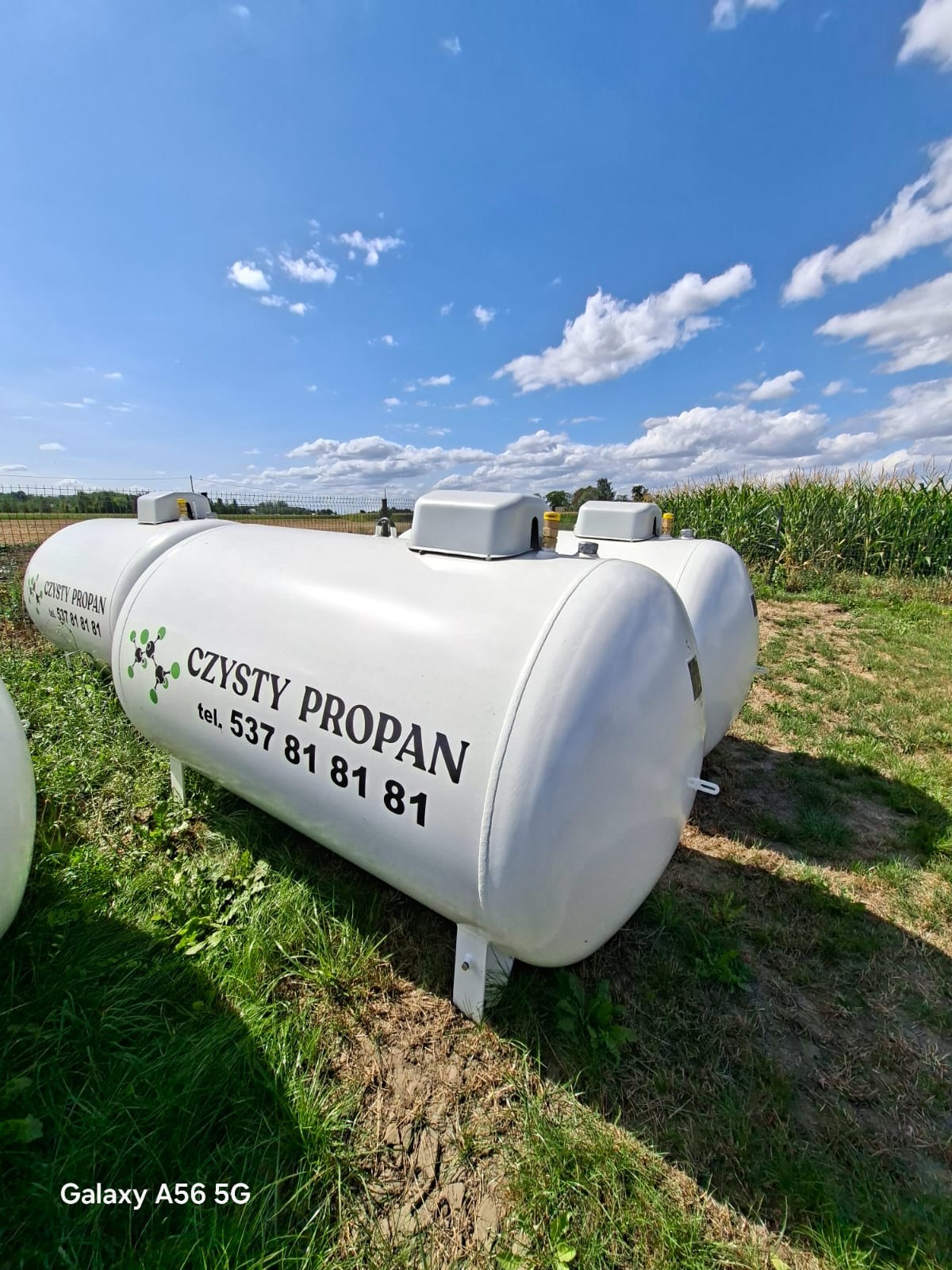 Białe zbiorniki na propan z napisem 'Czysty Propan' i numerem telefonu, ustawione na trawie pod błękitnym niebem z chmurami. Widok z dołu, w tle pole kukurydzy.
