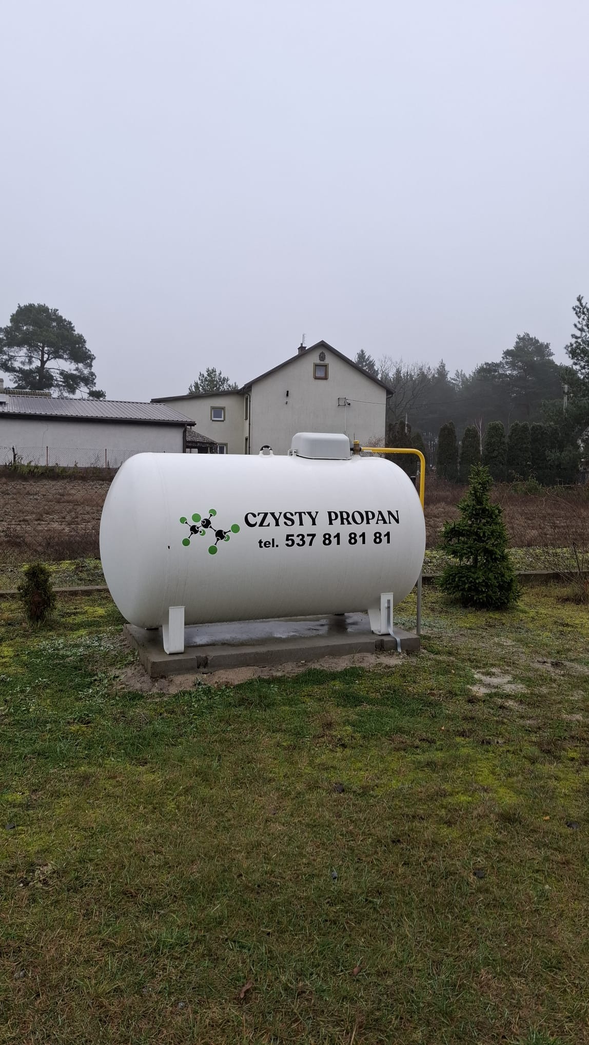 Biały zbiornik na gaz propan z napisem 'CZYSTY PROPAN' i numerem telefonu, umieszczony na betonowej podstawie na zewnątrz, na tle domu i drzew.