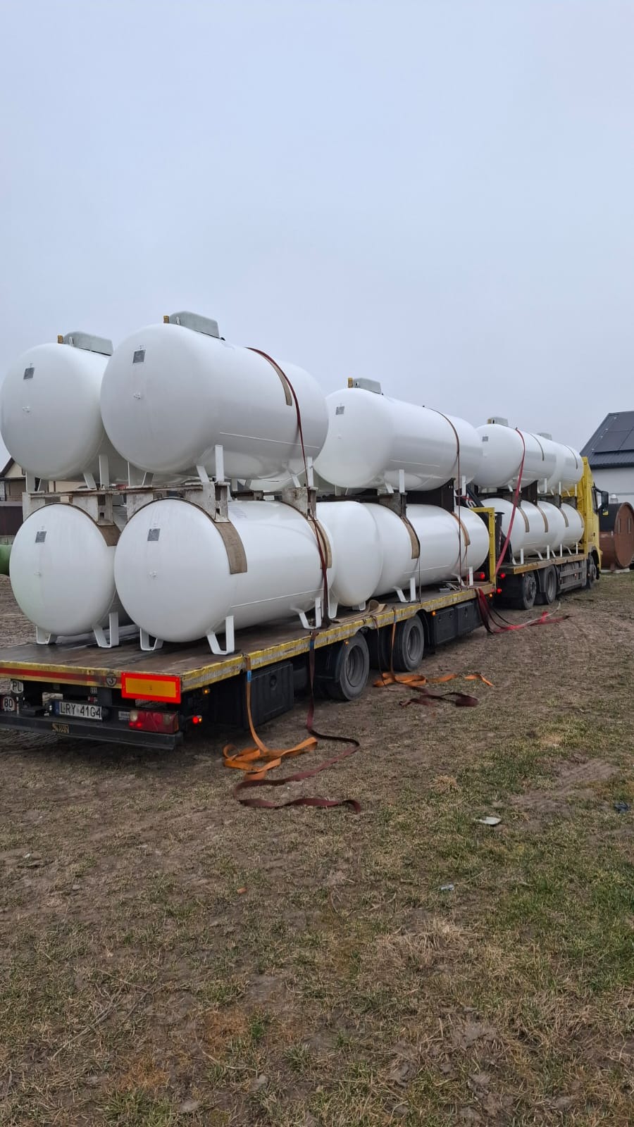 Transport białych zbiorników gazowych na naczepie ciężarówki, zabezpieczonych pasami. Widok z tyłu pojazdu na tle zabudowań.