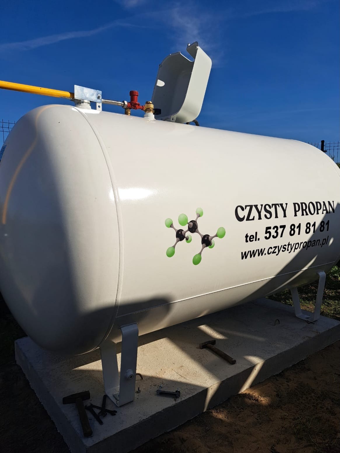 Biały zbiornik na propan z żółtym przyłączem, umieszczony na betonowej podstawie. Widoczne narzędzia i logo 'Czysty Propan' z numerem telefonu i adresem strony.