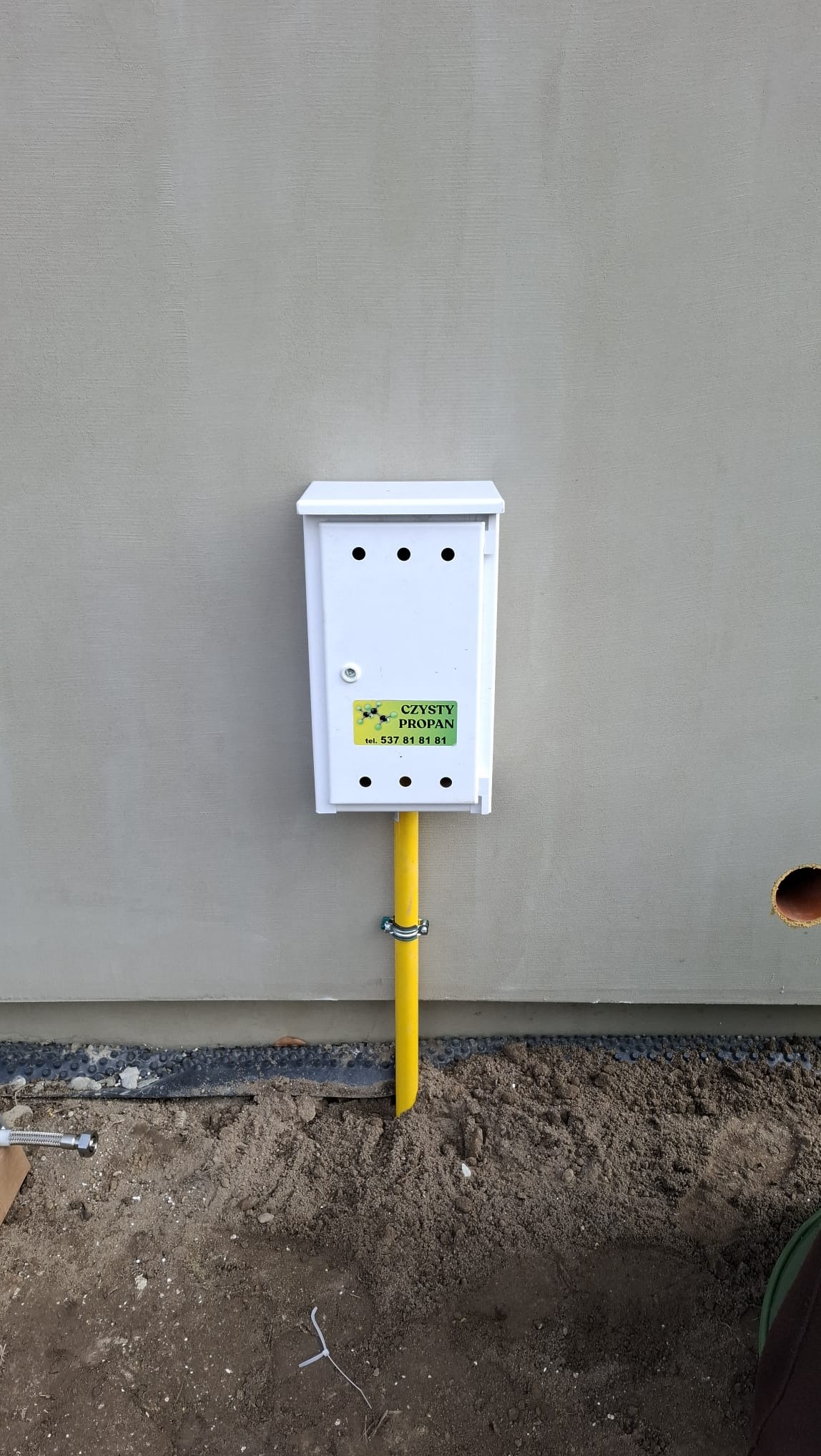 Biała skrzynka gazowa z żółtą rurą przy ścianie budynku, z naklejką 'Czysty Propan' i numerem telefonu. Widoczny fragment gruntu i otwór w ścianie.
