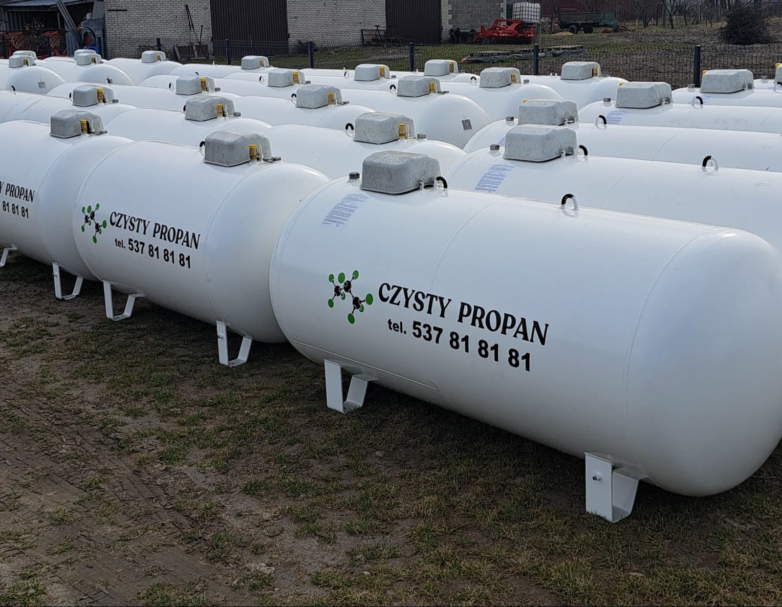 Białe zbiorniki na propan z napisem 'Czysty Propan' i numerem telefonu, ustawione rzędami na zewnątrz, z budynkiem i drzewami w tle. Widok z perspektywy.