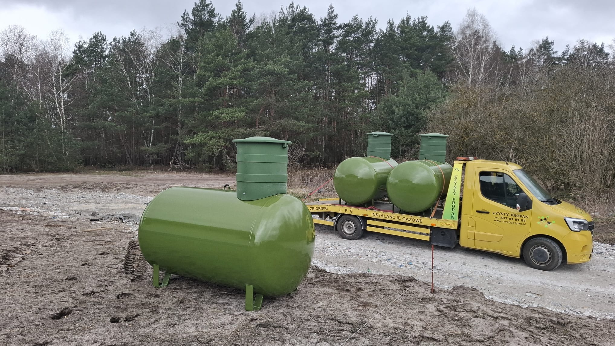 Zielony zbiornik LPG na ziemi, w tle przyczepa z kolejnymi zbiornikami i żółty samochód dostawczy firmy Czysty Propan. Las w tle.