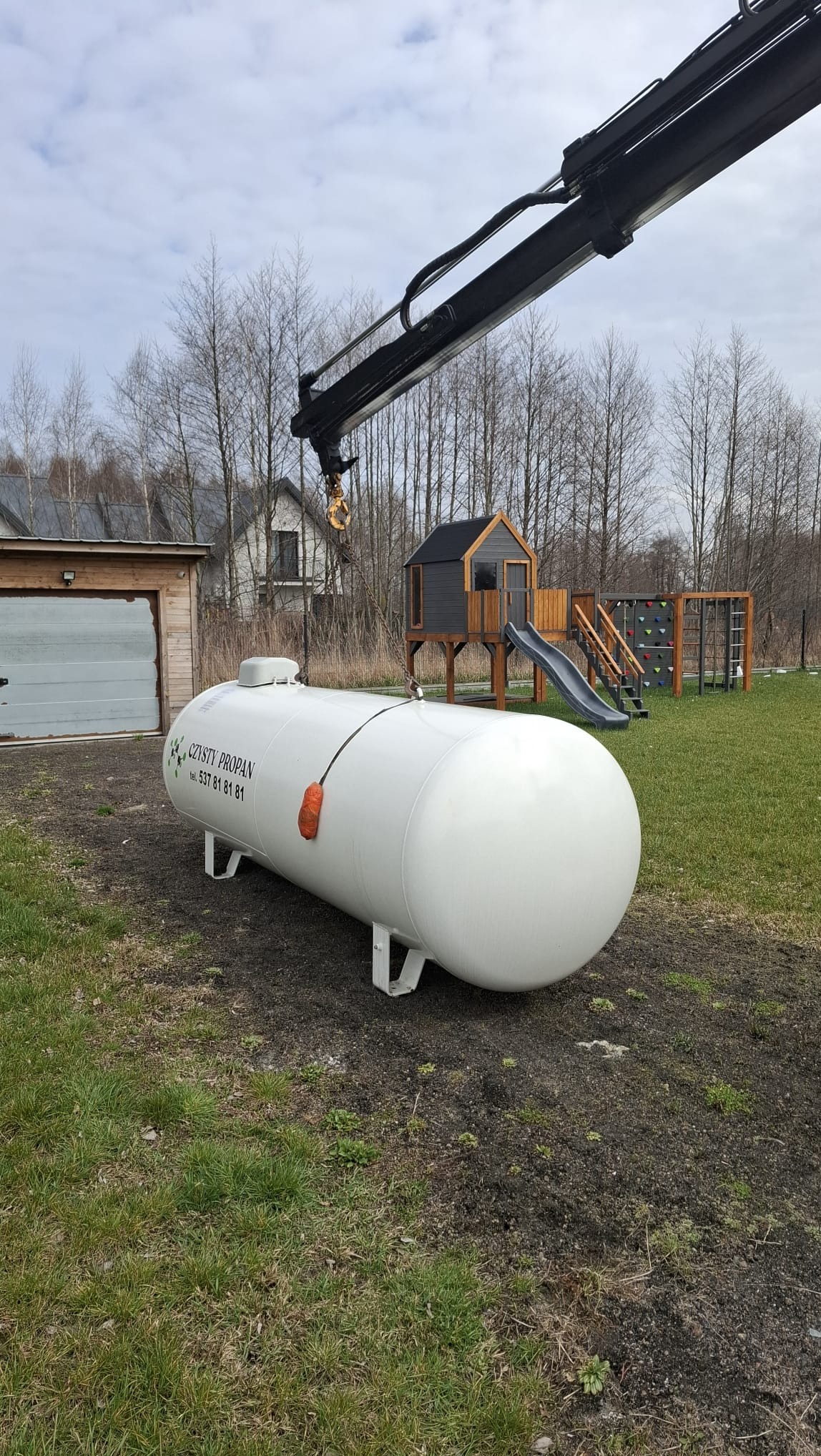 Biały zbiornik na gaz propan, umieszczony na trawie, podnoszony przez ramię dźwigu. W tle budynek gospodarczy i plac zabaw.