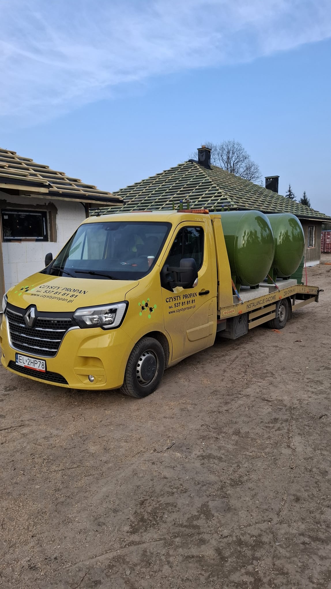 Żółty samochód dostawczy z dwoma zielonymi zbiornikami LPG na platformie, na tle budynków w trakcie budowy. Widoczne logo firmy 'Czysty Propan'.