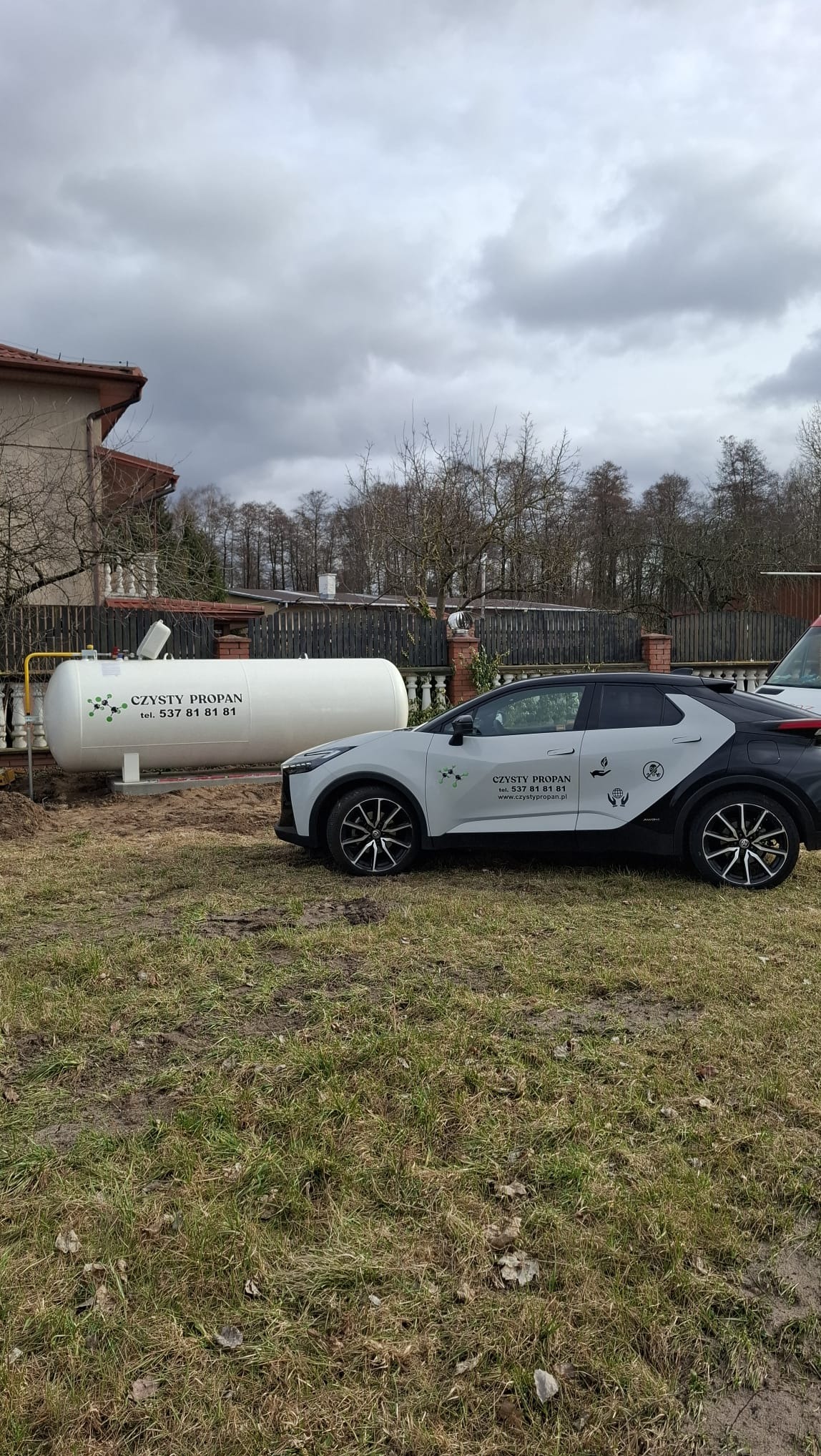 Biały zbiornik na gaz propan z żółtym przyłączem, obok zaparkowany samochód z logo firmy. Pochmurne niebo w tle.