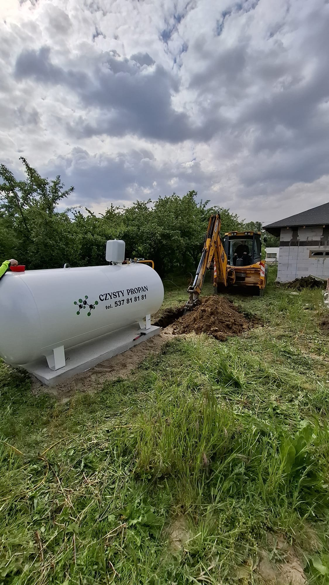 Biały zbiornik na gaz propan z napisem i numerem telefonu, umieszczony na betonowej podstawie. W tle koparka przygotowuje wykop, a dalej widać budynek w budowie.