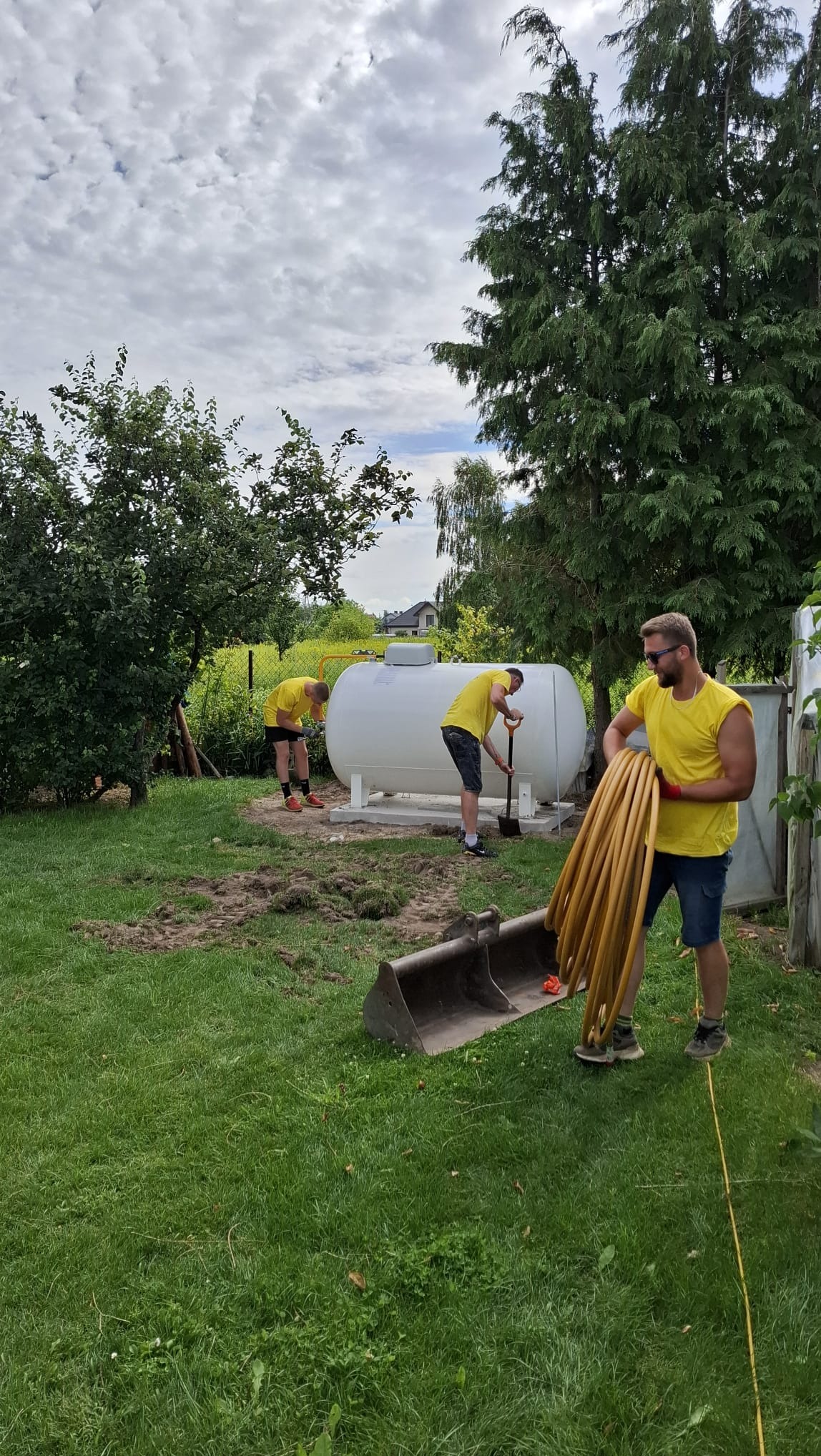 Montaż zbiornika na gaz płynny w ogrodzie: trzech mężczyzn w żółtych koszulkach pracuje przy białym zbiorniku, jeden kopie, drugi trzyma rury, w tle zieleń i błękitne niebo.
