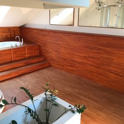 GLAZART - Glazurnictwo Remonty Wykończenia - Drewnem wykończona łazienka na poddaszu z wanną na podeście i kabiną prysznicową widoczną w lustrze. Parkiet na podłodze i roślina w donicy na pierwszym planie.