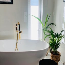 GLAZART - Glazurnictwo Remonty Wykończenia - Nowoczesna łazienka z białą wanną wolnostojącą, złotą armaturą i roślinami doniczkowymi. Na ścianie grafika z mapą Warszawy. Minimalistyczny design.
