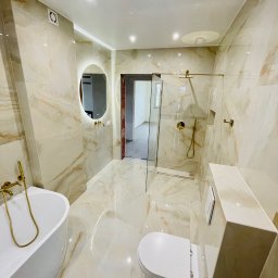 GLAZART - Glazurnictwo Remonty Wykończenia - Elegancka łazienka z beżowymi płytkami imitującymi marmur, złotą armaturą, owalnym lustrem z podświetleniem i kabiną prysznicową typu walk-in.
