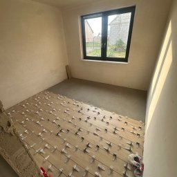 GLAZART - Glazurnictwo Remonty Wykończenia - Pokój w trakcie układania drewnianej podłogi z użyciem klipsów poziomujących. Widoczne surowe ściany i okno z widokiem na zewnątrz. Częściowo ułożona podłoga.