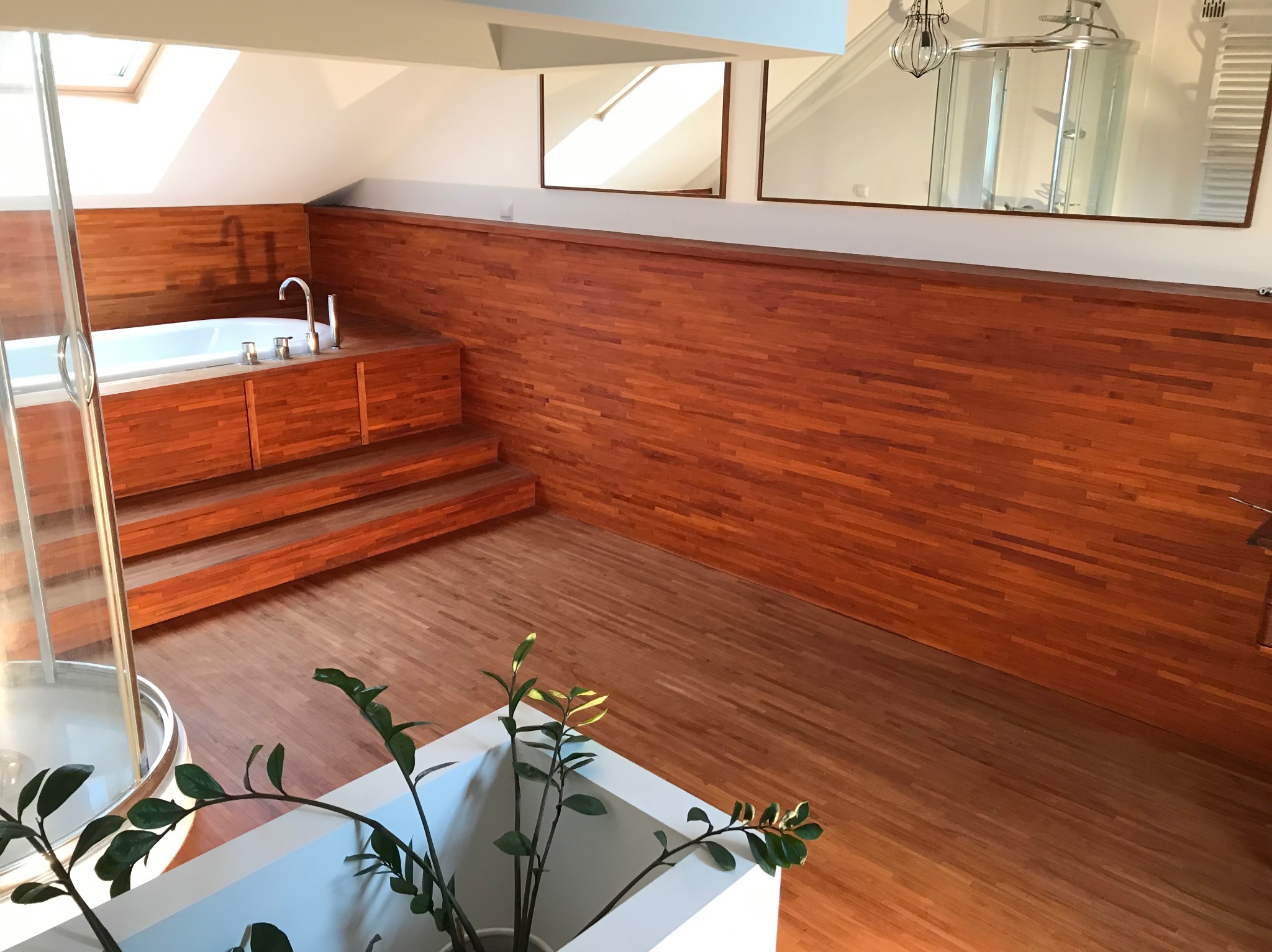 Drewnem wykończona łazienka na poddaszu z wanną na podeście i kabiną prysznicową widoczną w lustrze. Parkiet na podłodze i roślina w donicy na pierwszym planie.