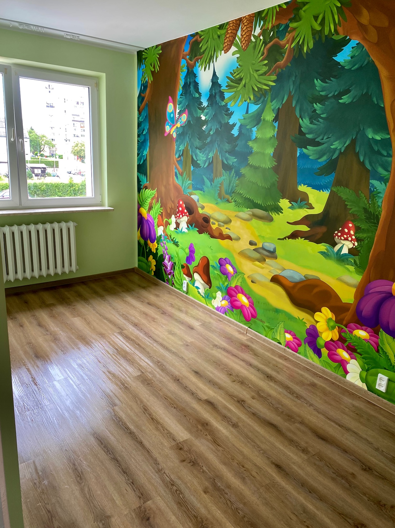 Pokój dziecięcy z laminowaną podłogą i ścianą ozdobioną malowidłem leśnym z grzybami, kwiatami i motylem. Okno z grzejnikiem po lewej stronie.