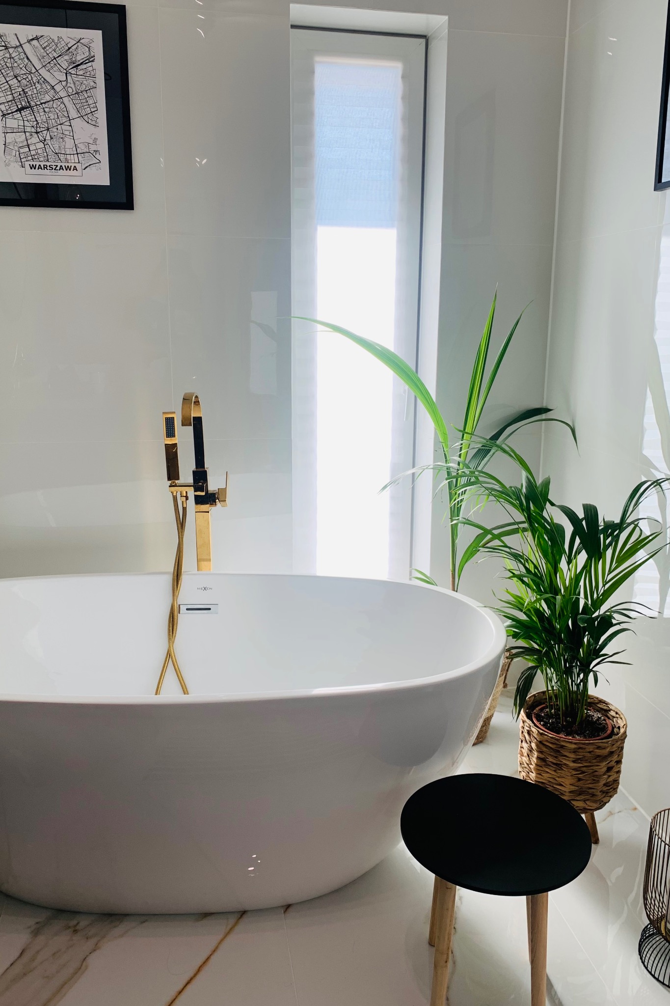 Nowoczesna łazienka z białą wanną wolnostojącą, złotą armaturą i roślinami doniczkowymi. Na ścianie grafika z mapą Warszawy. Minimalistyczny design.