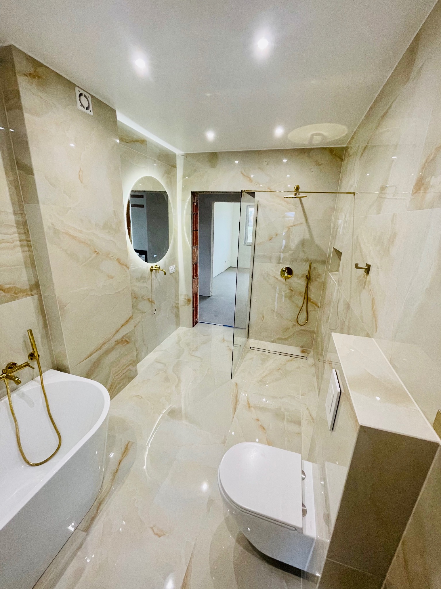 Elegancka łazienka z beżowymi płytkami imitującymi marmur, złotą armaturą, owalnym lustrem z podświetleniem i kabiną prysznicową typu walk-in.