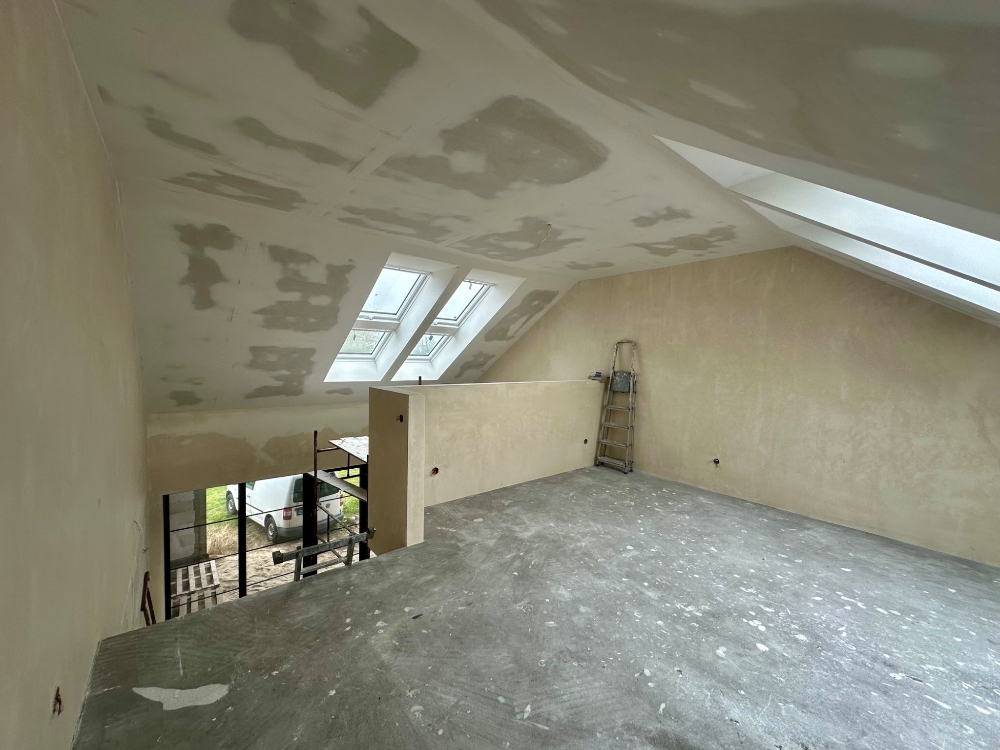 Poddasze w trakcie prac wykończeniowych: szpachlowane ściany i sufit z oknami dachowymi, widoczna drabina i rusztowanie. Surowa podłoga z plamami farby.