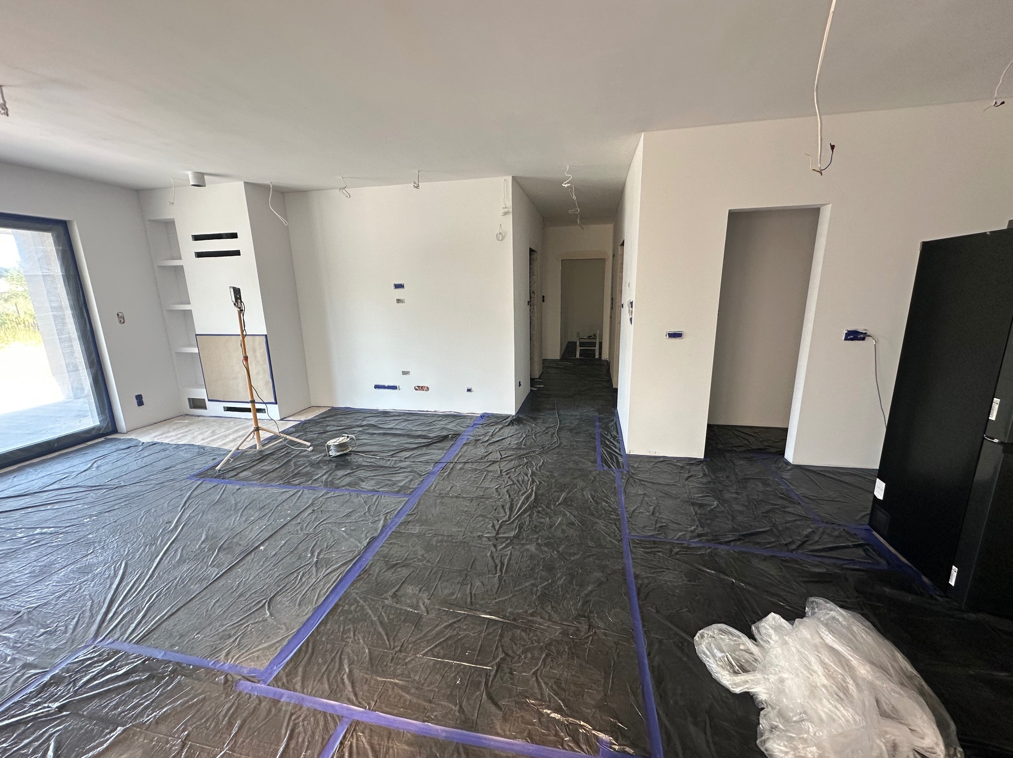 Przygotowanie do malowania: podłoga zabezpieczona folią malarską z niebieską taśmą, ściany świeżo pomalowane na biało, widoczne kable i lampa na statywie.