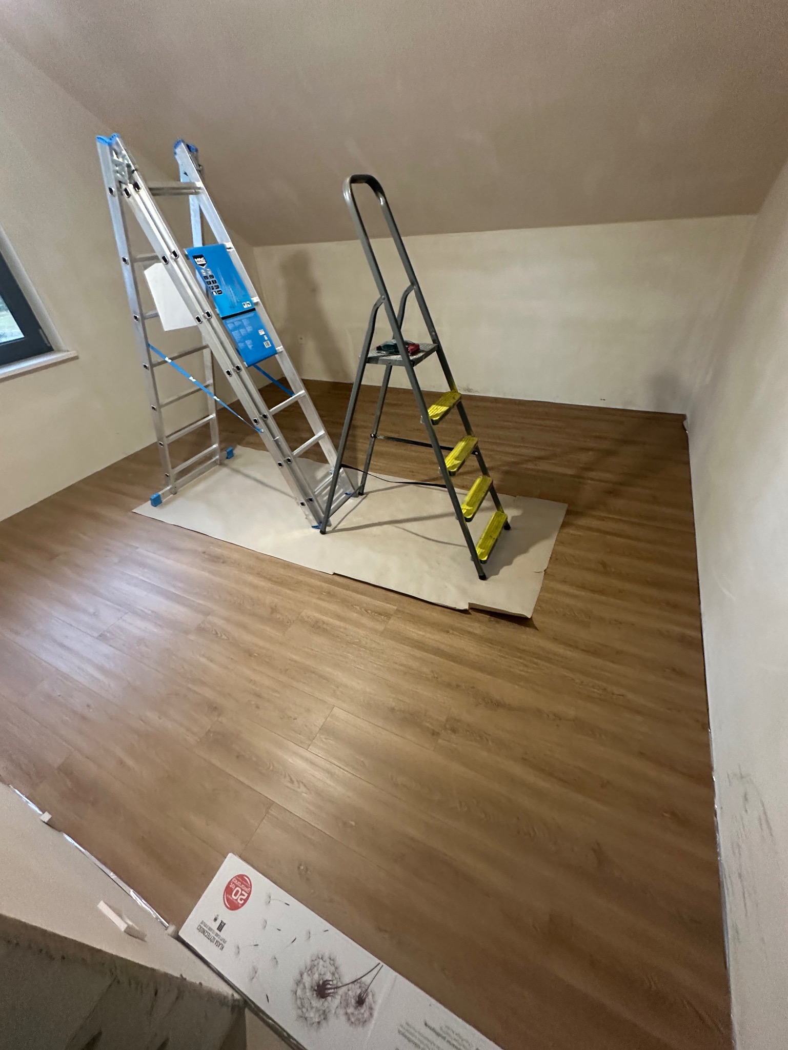 Poddasze z ułożoną podłogą z paneli, dwie drabiny rozstawione na kartonie ochronnym, w tle ściany przygotowane do malowania, opakowanie paneli pod ścianą.
