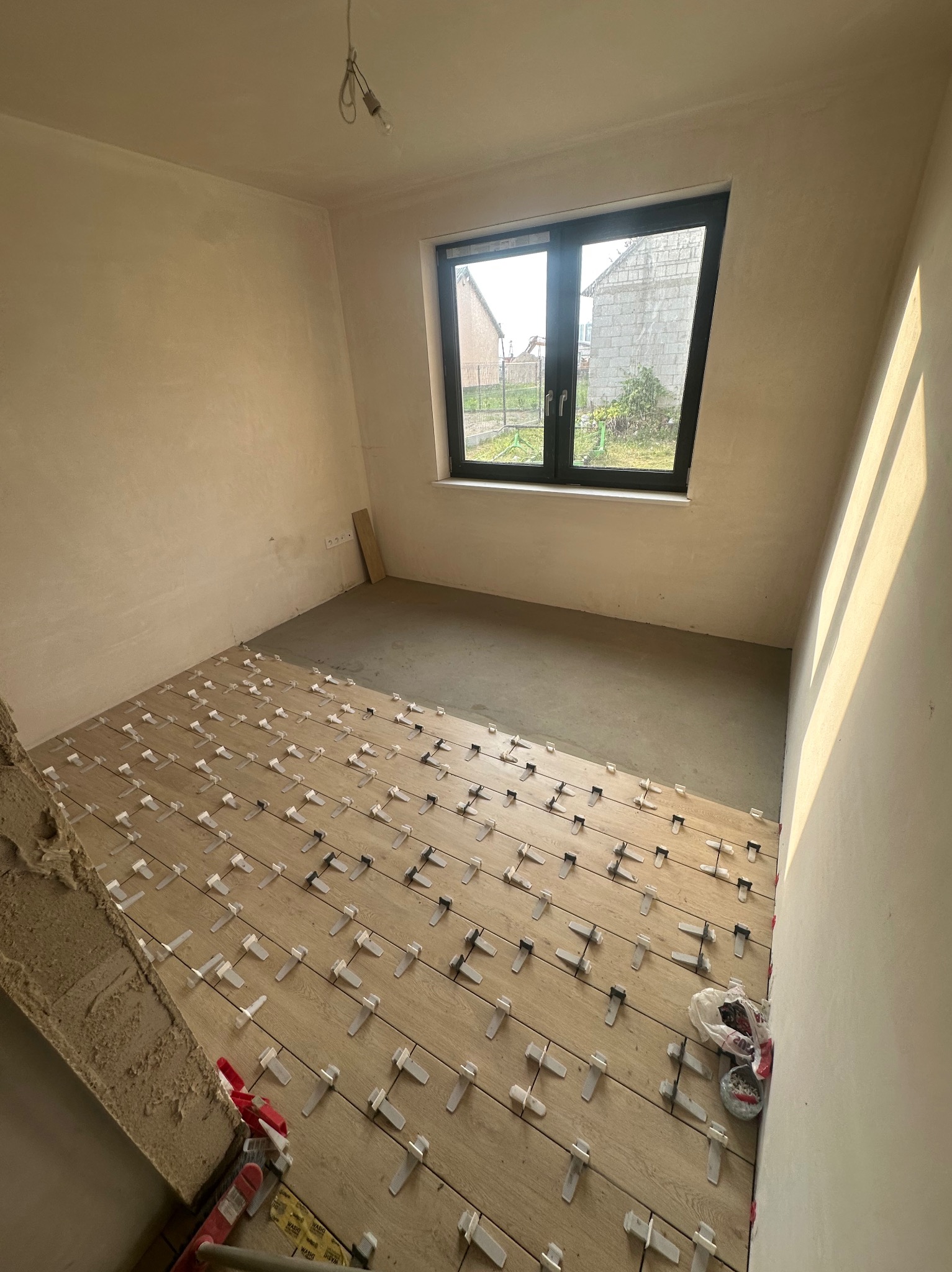 Pokój w trakcie układania drewnianej podłogi z użyciem klipsów poziomujących. Widoczne surowe ściany i okno z widokiem na zewnątrz. Częściowo ułożona podłoga.