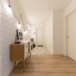 A-PROject Alexandr Karpeliov - Nowoczesny przedpokój z białą cegłą, drewnianą podłogą i komodą w stylu retro. Dekoracyjne oświetlenie żarówkowe i geometryczna tapeta dodają wnętrzu charakteru.