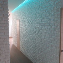 A-PROject Alexandr Karpeliov - Biała ściana z cegły dekoracyjnej, oświetlona niebieskim światłem LED, tworząca nowoczesny wygląd korytarza z drewnianą podłogą i ukrytymi drzwiami.