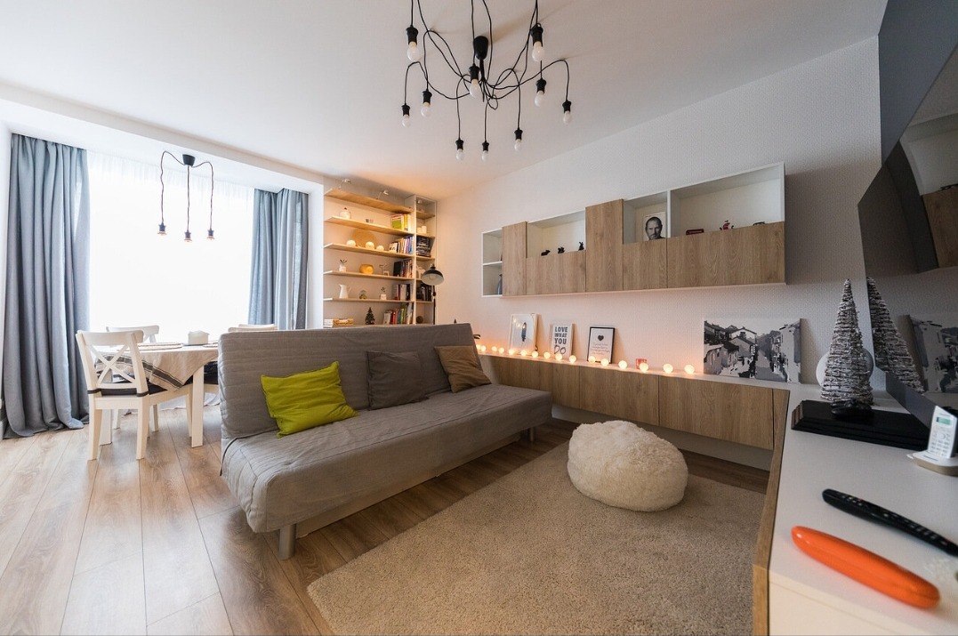 Jasne wnętrze pokoju dziennego z drewnianą podłogą, szarą sofą z zieloną poduszką i regałem z książkami w tle. Dekoracyjne oświetlenie dodaje przytulności.