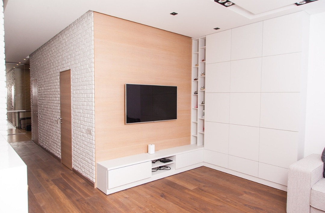 Nowoczesny salon z ceglaną ścianą, drewnianą podłogą i minimalistyczną zabudową meblową w bieli. Telewizor na ścianie, subtelne oświetlenie.