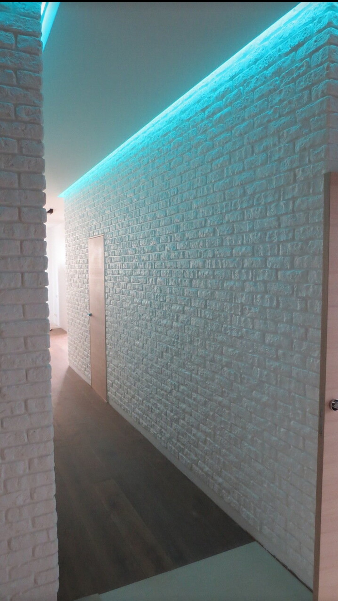Biała ściana z cegły dekoracyjnej, oświetlona niebieskim światłem LED, tworząca nowoczesny wygląd korytarza z drewnianą podłogą i ukrytymi drzwiami.