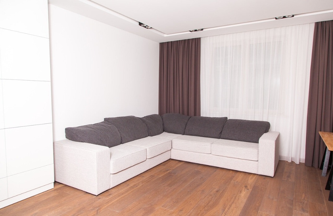 Jasny narożnik z szarymi poduszkami w rogu pokoju z drewnianą podłogą. Minimalistyczny design, brązowe zasłony i białe ściany. Nowoczesne oświetlenie sufitowe.