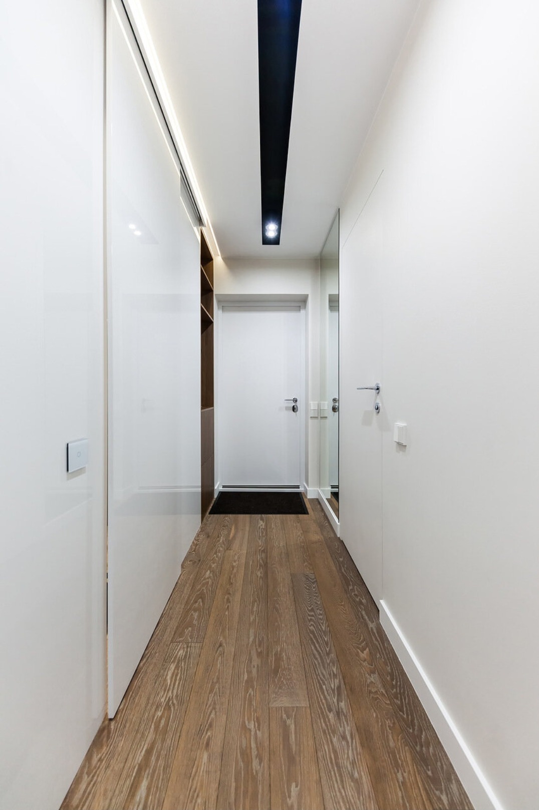 Nowoczesny korytarz z drewnianą podłogą, białymi ścianami i przesuwnymi drzwiami. Długi, wąski korytarz z oświetleniem sufitowym i minimalistycznym designem.