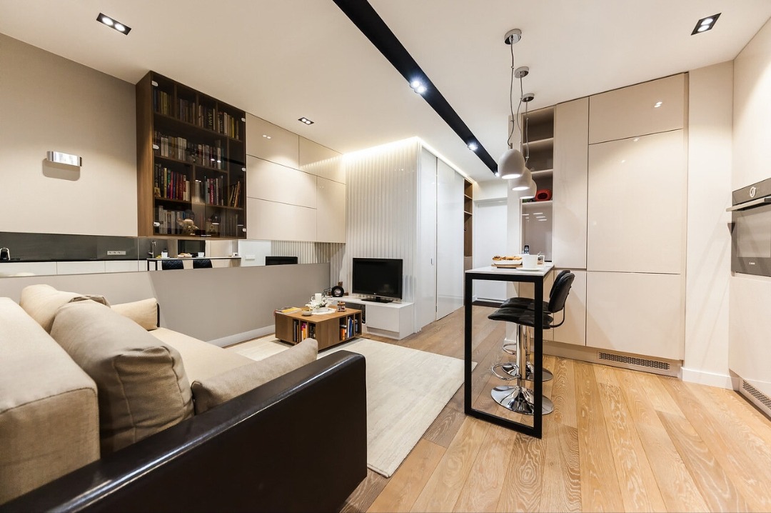 Nowoczesne wnętrze salonu z aneksem kuchennym: beżowa sofa, drewniana podłoga, minimalistyczny stół barowy i zabudowa meblowa w jasnych odcieniach.