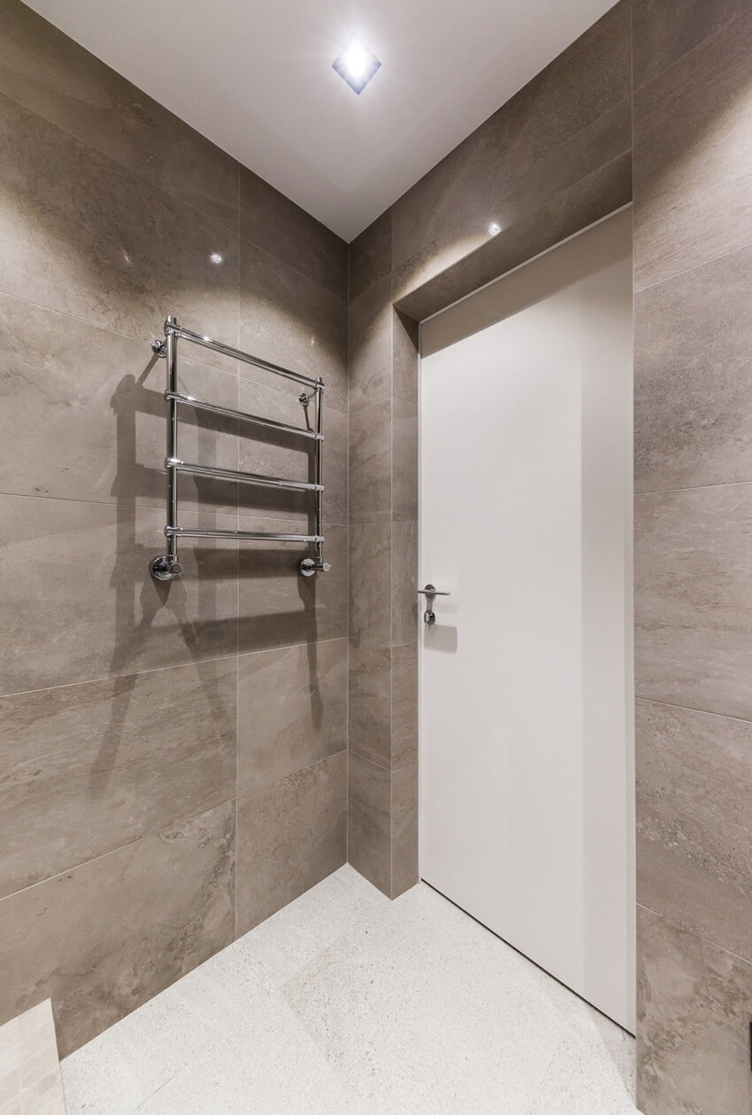 Wykończona łazienka w Warszawie: beżowe płytki imitujące kamień na ścianach, biała podłoga z drobnymi kamyczkami, chromowany grzejnik drabinkowy i białe drzwi.