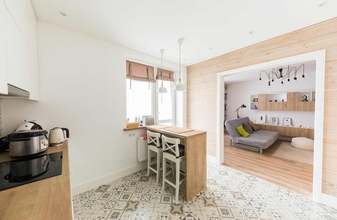 Jasna kuchnia z drewnianym blatem i wzorzystą podłogą. W tle widoczny salon z drewnianą ścianą i szarą sofą. Całość utrzymana w nowoczesnym, skandynawskim stylu.