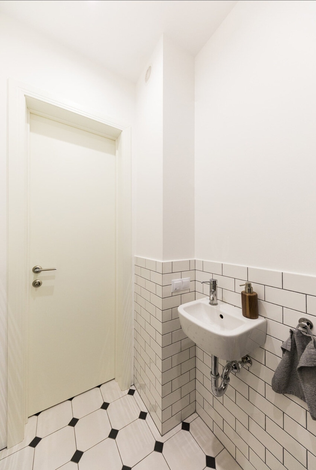 Biała, mała łazienka z umywalką, baterią i dozownikiem mydła. Ściany wyłożone białymi, prostokątnymi płytkami. Na podłodze biało-czarna mozaika. Widoczna biała rama drzwi.