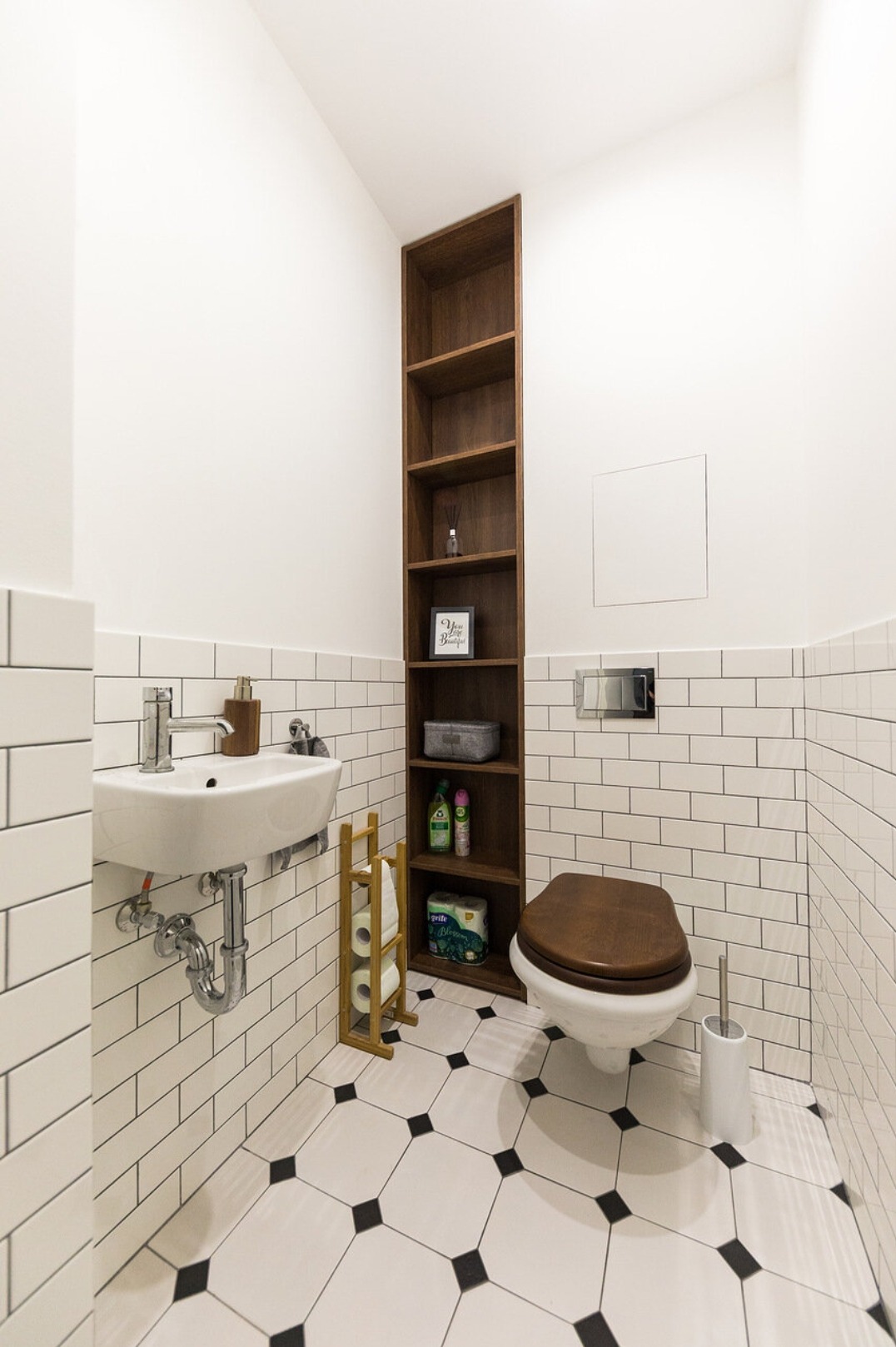 Biała łazienka z czarnymi akcentami: umywalka, toaleta z drewnianą deską, wysoka półka w zabudowie. Minimalistyczny design, płytki imitujące cegłę, wzorzysta podłoga.