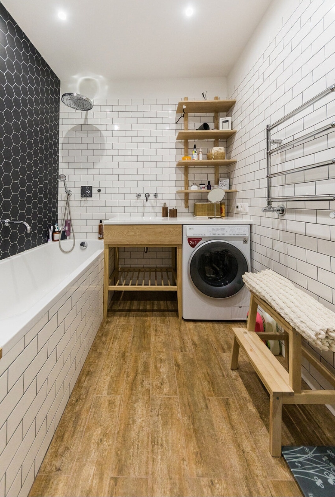 Nowoczesna łazienka z wanną, heksagonalną mozaiką, drewnianymi półkami i podłogą imitującą drewno. Pralka wkomponowana w zabudowę. Jasne, ciepłe oświetlenie.