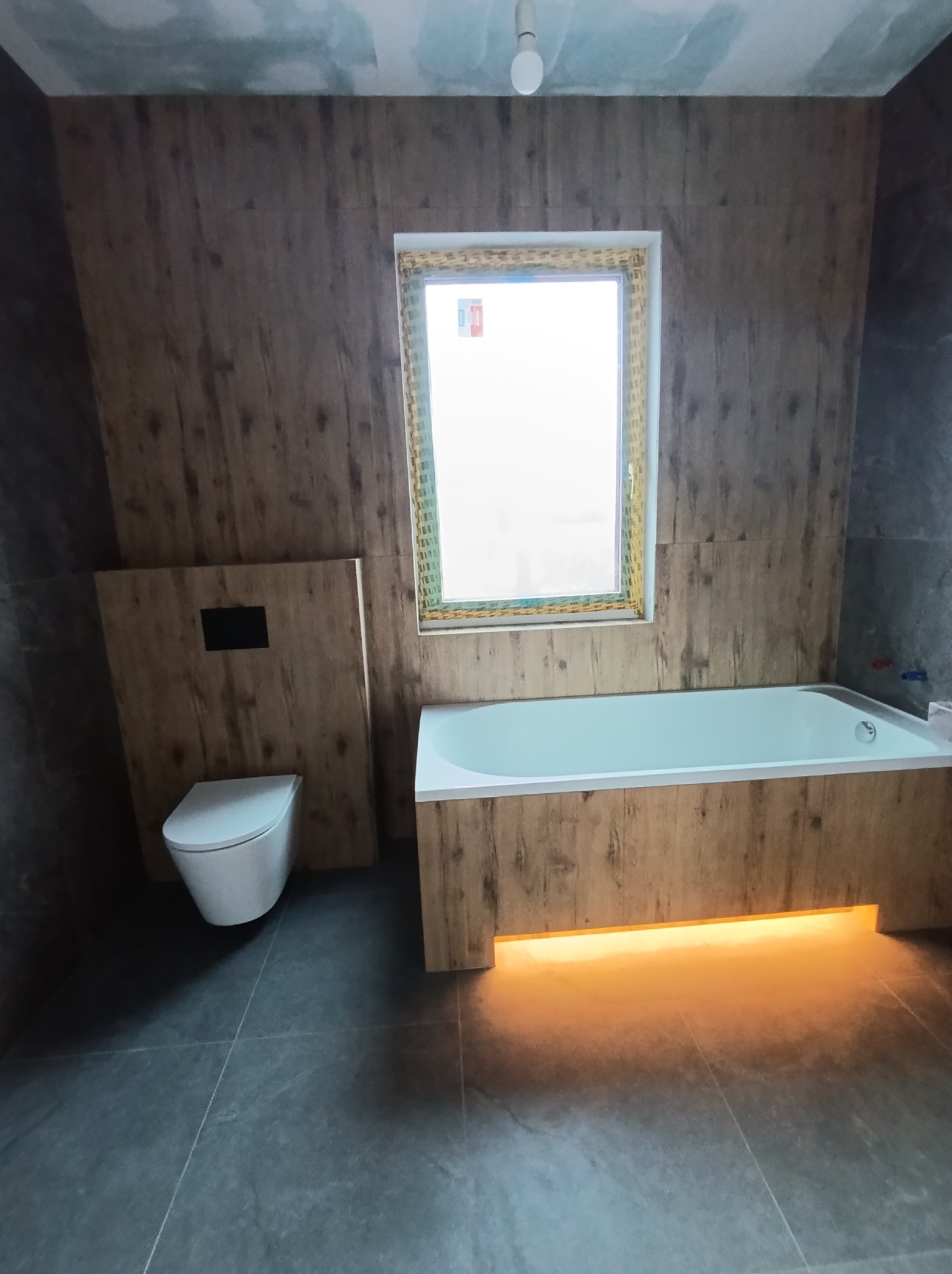 Nowoczesna łazienka z drewnopodobnymi płytkami, podświetlaną wanną i podwieszaną toaletą. Minimalistyczny design z akcentem na naturalne materiały i ciepłe oświetlenie.