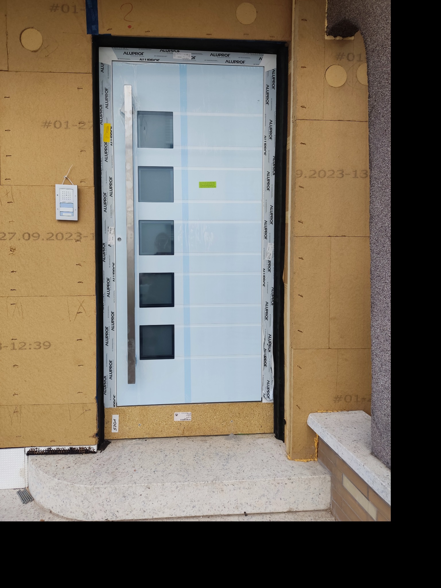 Białe drzwi wejściowe z czterema kwadratowymi przeszkleniami, zamontowane w surowym otworze, z widoczną izolacją budynku i elementami wykończeniowymi.