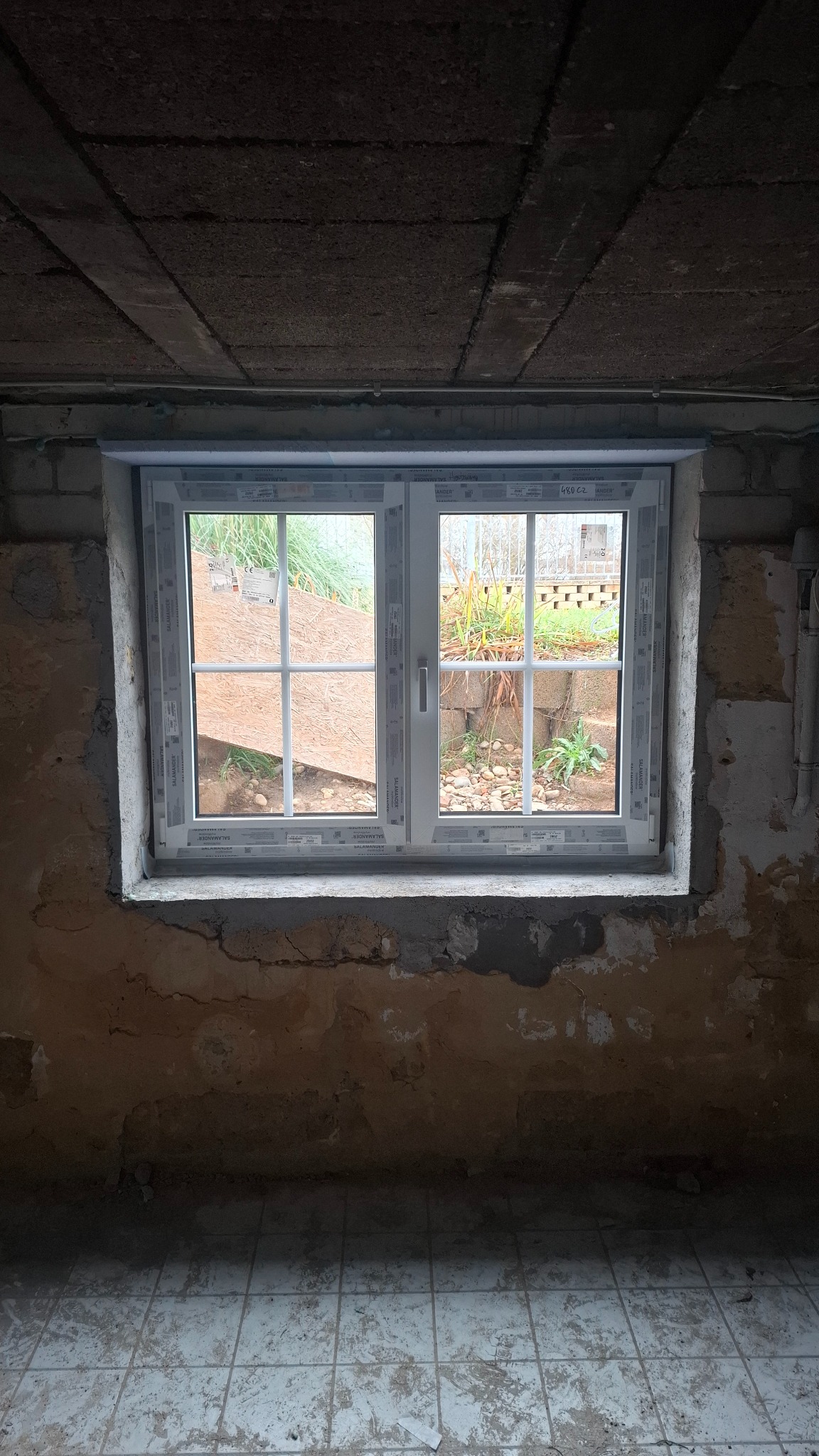 Białe, dwuskrzydłowe okno z kratką, zamontowane w surowym wnętrzu z widoczną cegłą i betonowym sufitem. Za oknem widok na ogród z roślinnością i elementami architektury.