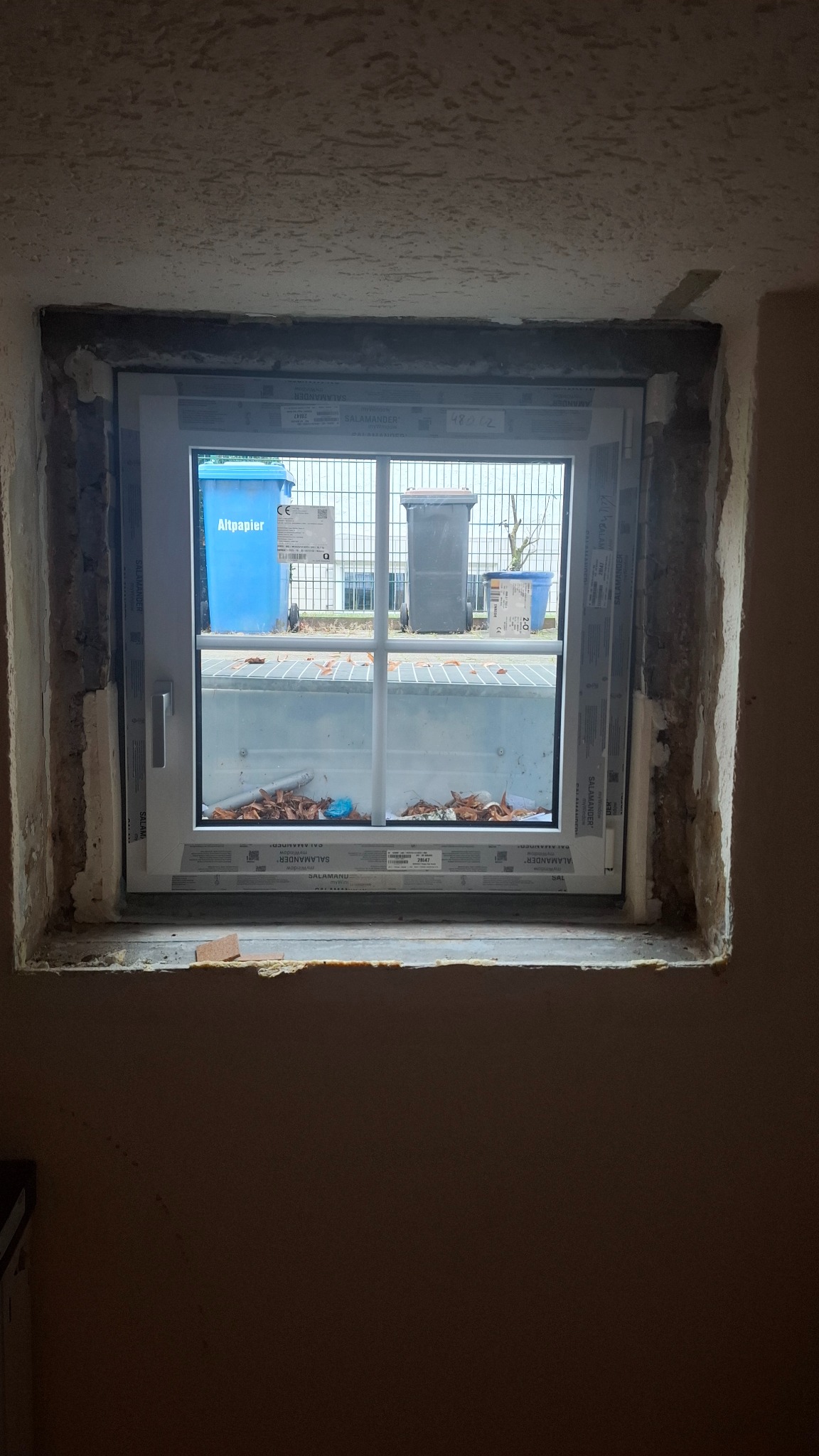 Montaż okna PCV Salamander w surowym otworze okiennym. Widoczne ślady pianki montażowej i gruzu. Za oknem pojemniki na śmieci i metalowa krata.