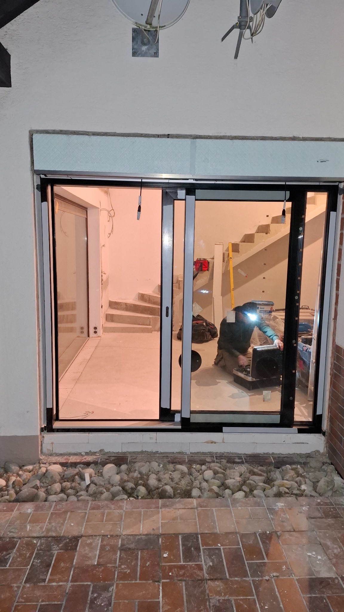 Montaż drzwi przesuwnych z czarną ramą. Widoczny pracownik w trakcie instalacji, narzędzia i schody w tle. Kamienie dekoracyjne u podstawy.