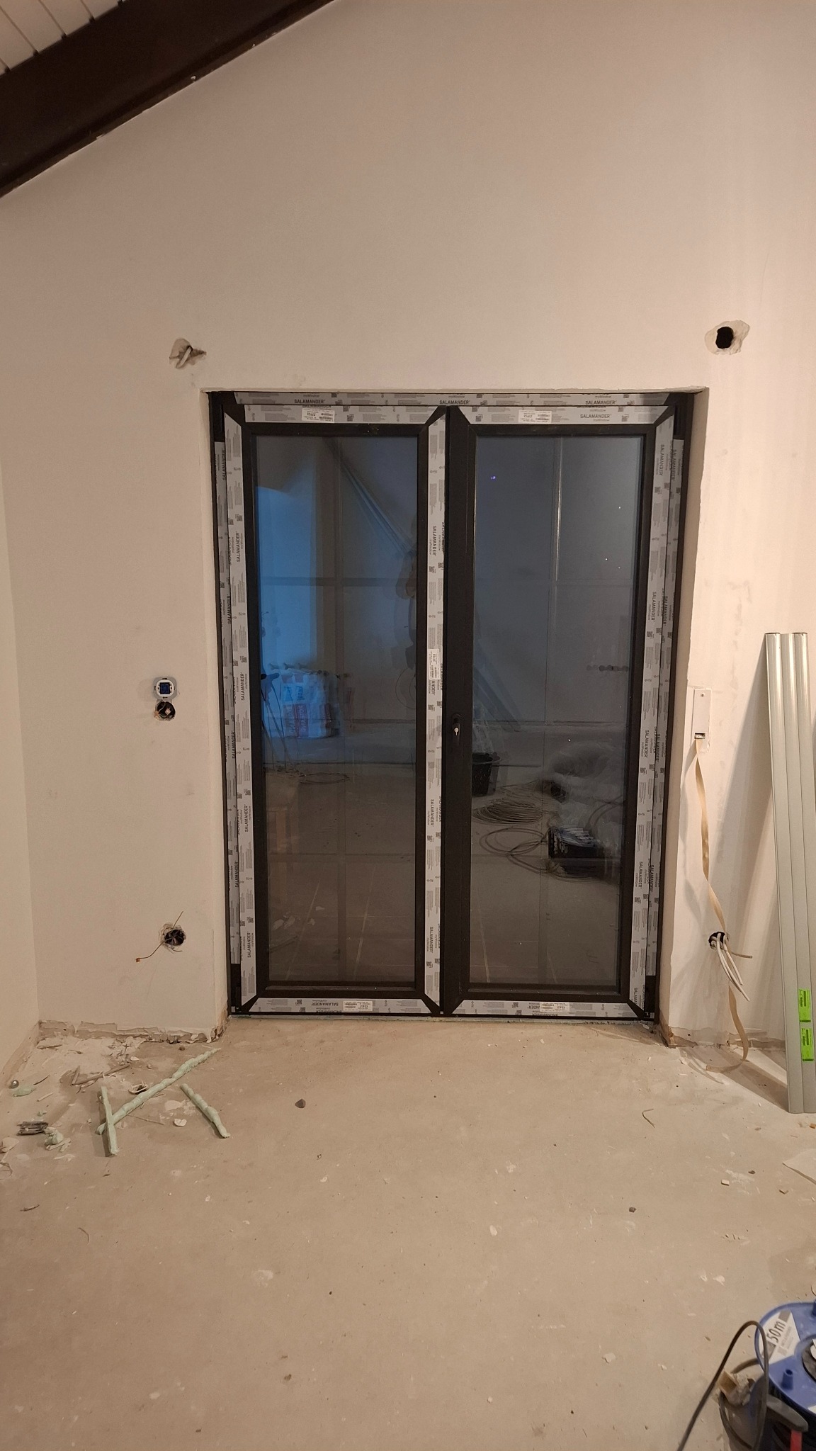 Zamontowane dwuskrzydłowe drzwi balkonowe z czarnymi ramami, widoczne ślady prac wykończeniowych na podłodze i ścianach. W tle widoczne materiały budowlane.