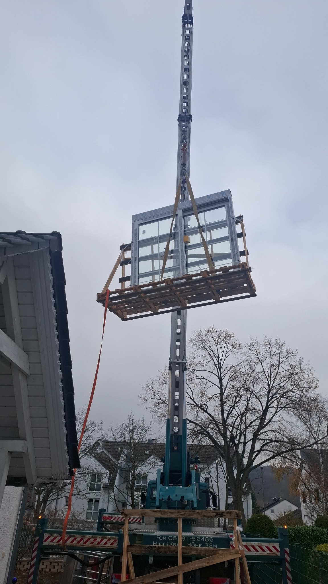 Montaż okna za pomocą dźwigu na tle pochmurnego nieba i zabudowy mieszkalnej. Widoczne drzewa bez liści i fragment dachu budynku.